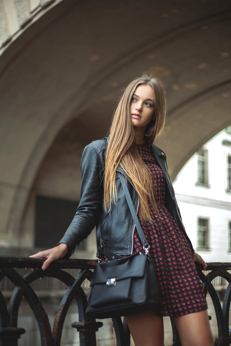 Stylish woman poses with a black leather jacket and dress in a city street, embodying modern fashion.