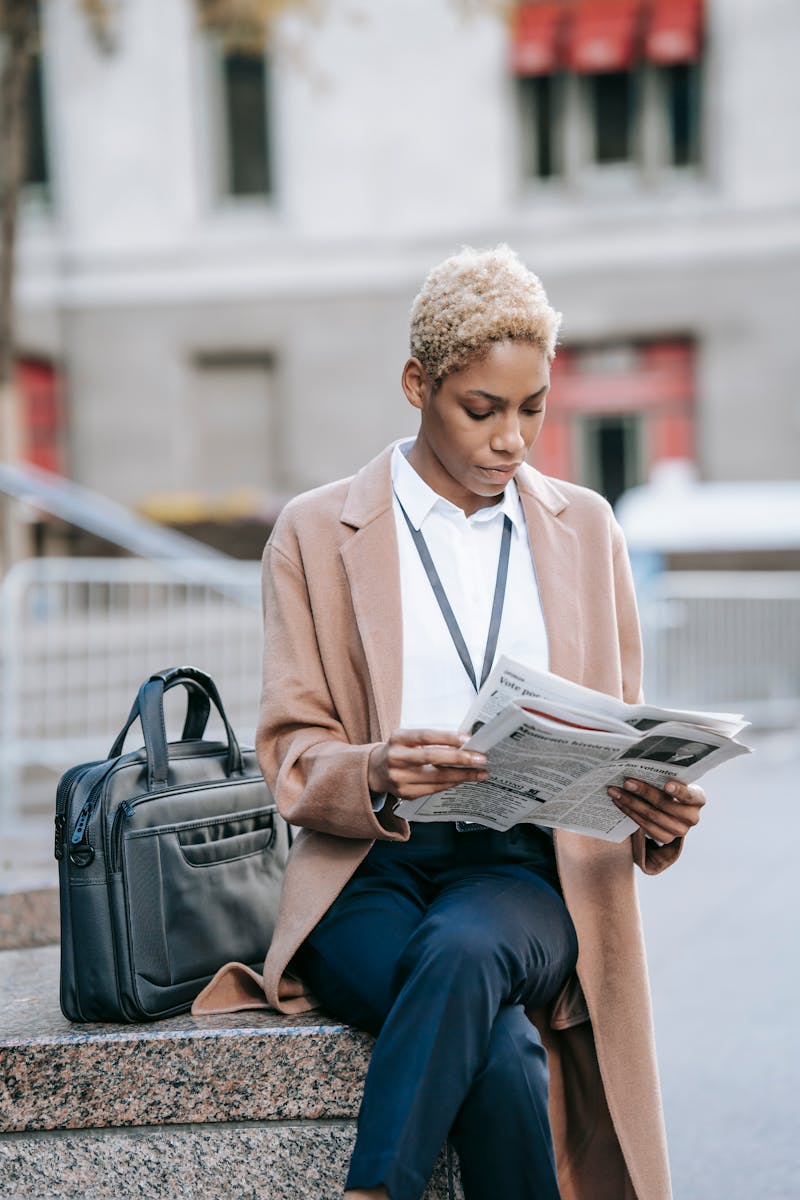Focused young African American female entrepreneur in elegant brown coat sitting on stone border near laptop bag with crossed legs and reading newspaper